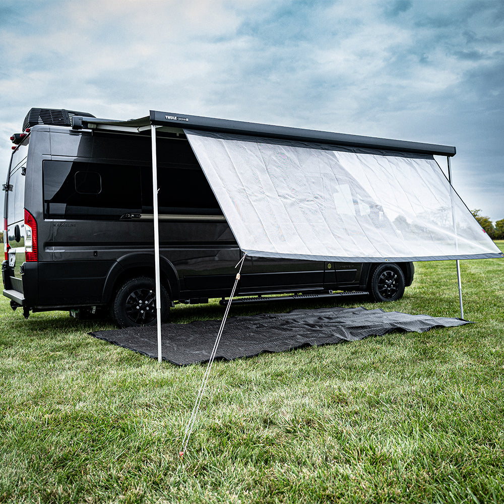 A black camper van with an extended white awning is parked on grass under a cloudy sky, featuring a Shade Screen for Airstream Rangeline for added sun protection. A black mat is laid out beneath the awning for outdoor use.