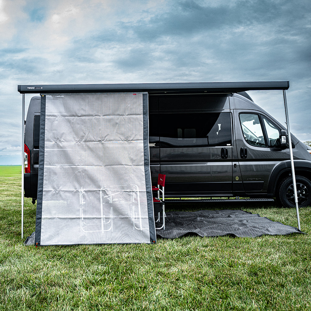 A black van is parked on grass with a Shade Screen for Airstream Rangeline in gray extended as an awning, and a mesh privacy screen attached for excellent sun protection. Behind the canopy shade screen, a red chair and a white metal rack are visible. The sky above is cloudy.