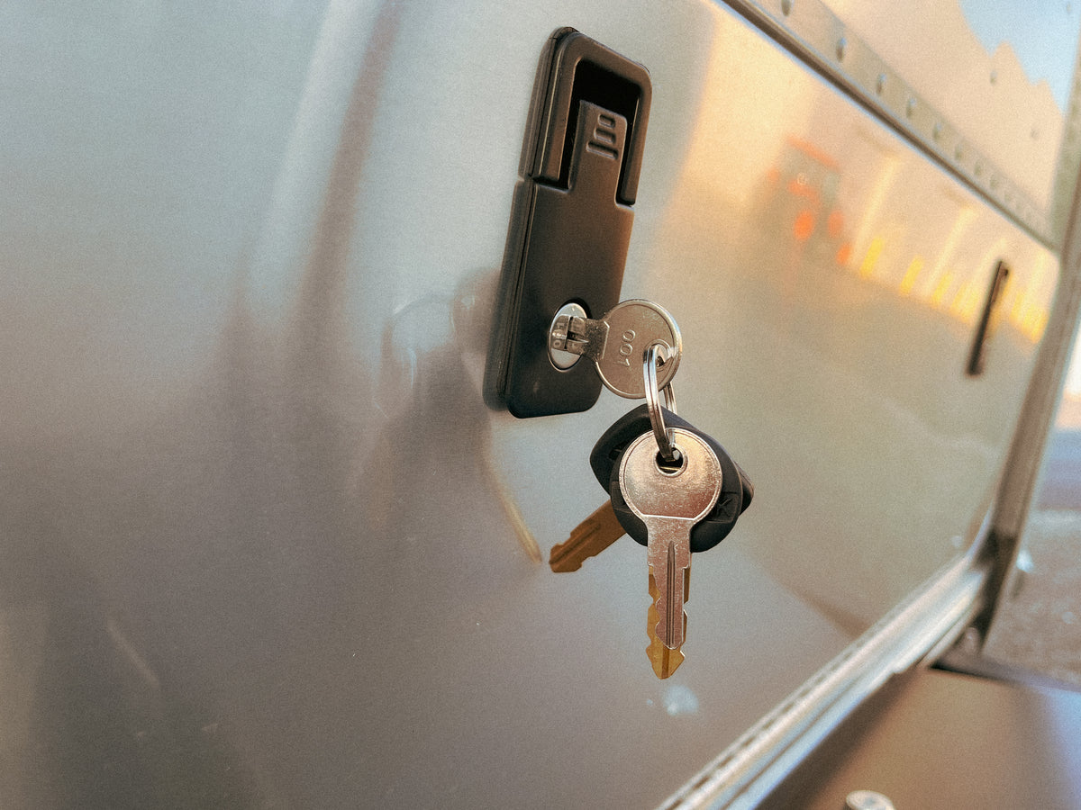 Close-up of two Airstream Compartment Door Pre-Cut Keys inserted into a lock on a metallic surface—likely part of an Airstream travel trailer or storage compartment—with subtle reflections visible on the polished metal.