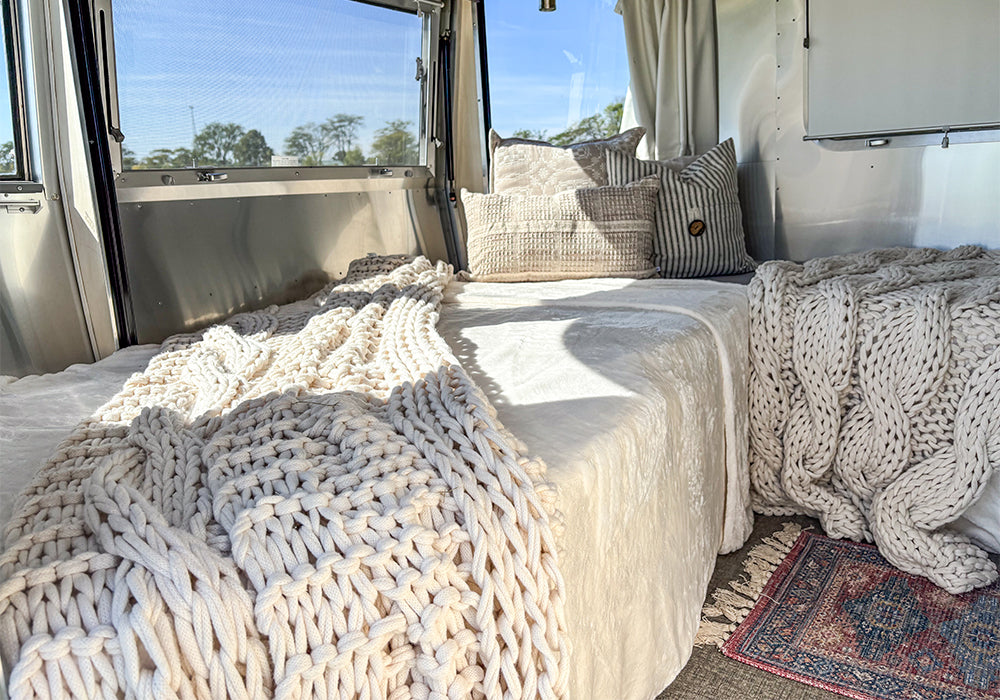 Relax in a sunlit Airstream surrounded by chunky knit blankets, textured pillows, and a small rug, while enjoying the view of trees outside—featuring the ultra-comfortable Airstream Sofa/Lounge Fitted Sheet for 28′ and 30′ International Travel Trailers for perfect bedding on the road.