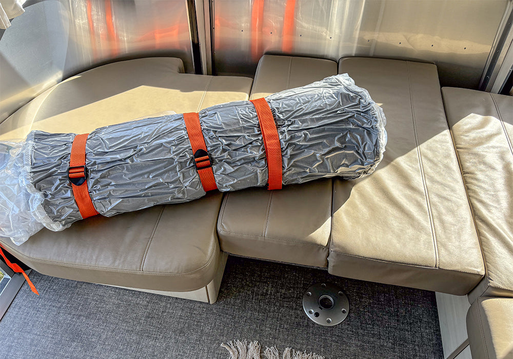 A rolled-up Airstream Dinette Memory Foam Topper for International Travel Trailers, wrapped in plastic and secured with orange straps, sits on tan cushioned seats inside a modern vehicle or trailer.