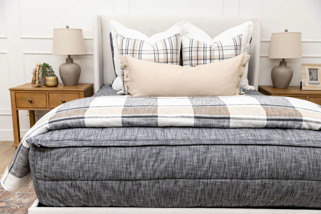 A cozy, neutral-toned bedroom features a neatly made bed with the Airstream RV Fit Beddy’s for Excella Travel Trailers, complemented by plaid and solid pillows, a striped comforter, and flanked by two wooden nightstands topped with lamps, a plant, and framed photos.