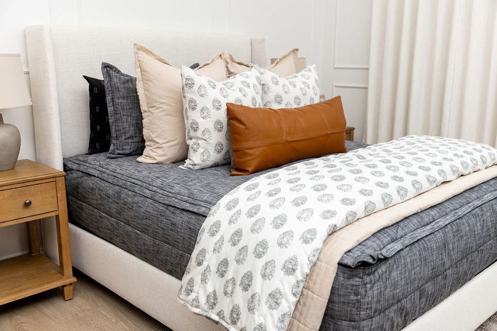 A neatly made bed featuring the Airstream RV Fit Beddy’s for Excella Travel Trailers, layered with pillows including a brown lumbar pillow, gray bedding, and a white patterned blanket creates a cozy retreat; this stylish all-in-one bedding set adds effortless comfort. A wooden nightstand with a lamp sits beside the bed.