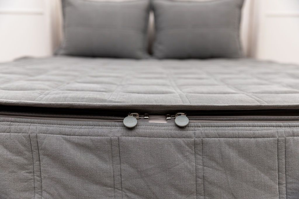 A close-up of a neatly made bed showcases the Airstream RV Fit Beddy’s for Flying Cloud Travel Trailers, featuring a gray quilted blanket with two convenient zippers at the foot, while two matching gray pillows are visible in the background.