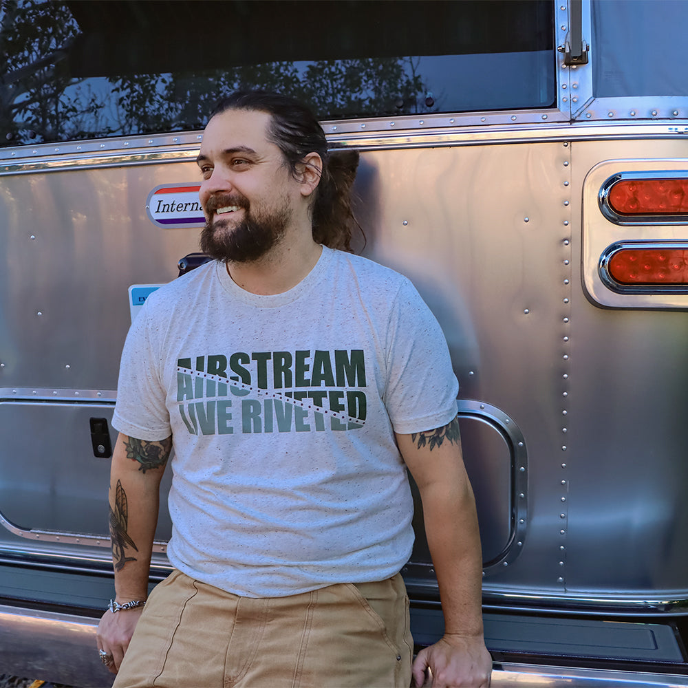 A bearded man with a ponytail smiles while sitting in front of an Airstream trailer. He wears the light gray Airstream Rivet Line T-Shirt, featuring “AIRSTREAM RIVETED” in bold green letters that proudly celebrate American craftsmanship, along with khaki pants.