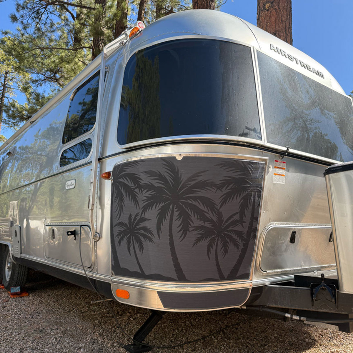 A silver Airstream trailer parked outdoors among trees, featuring a decorative panel with black silhouettes of palm trees on its front side.