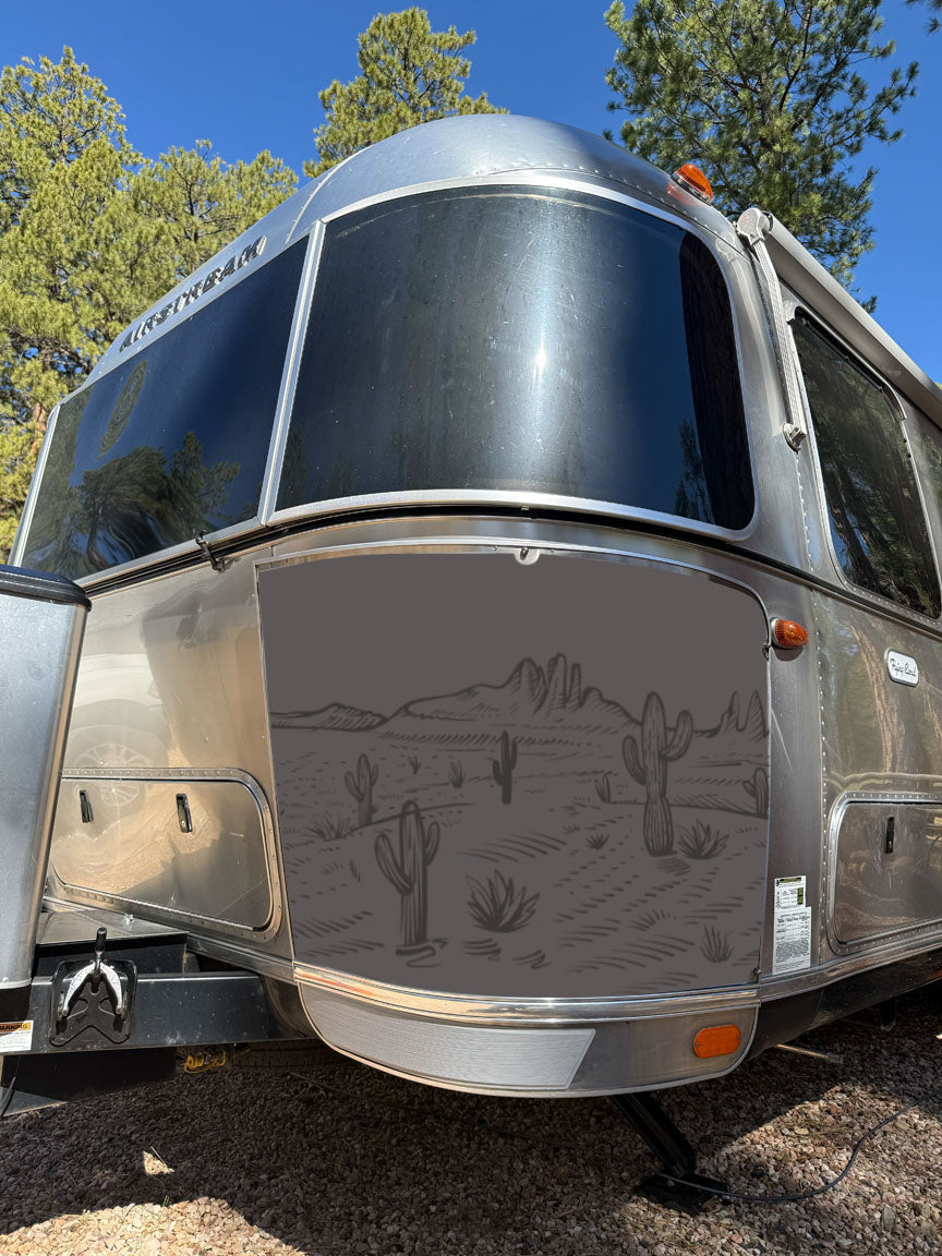 Airstream Rock Guard Rock Wraps Nature Collection: This silver Airstream trailer features a vibrant desert scene with cacti and mountains illustrated on its front, enhanced by durable marine-grade foam wraps for ultimate travel trailer protection. Pine trees stand in the sunny background, completing the nature-inspired design.