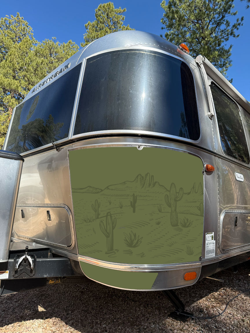 A silver RV featuring the Airstream Rock Guard Rock Wraps Nature Collection—with a desert landscape illustration of cacti and mountains on its front panel for enhanced travel trailer protection—is parked outdoors near pine trees beneath a clear blue sky.