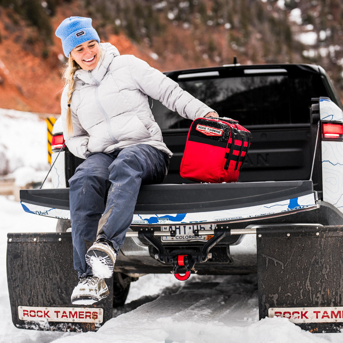 A smiling person dressed in winter clothes sits on a truck tailgate surrounded by snow and trees, holding a red bag. The truck is equipped with ROCK TAMERS mud flaps, while the Rock Tamers Recovery Rope Kit is conveniently nearby—an essential tool for off-road recovery in these snowy conditions.