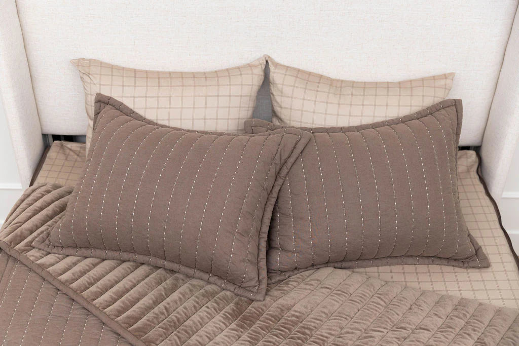Two brown quilted pillows and a matching Airstream RV Fit Beddy’s for Excella Travel Trailers rest on a beige bed with two beige checkered pillows propped behind them against a light-colored headboard, creating the perfect cozy setup for stylish Airstream bedding.