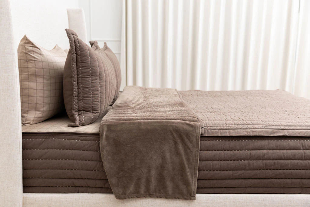 A neatly made bed features Airstream RV Fit Beddy’s for Pan America Travel Trailers in a rich brown quilted design with matching pillows, situated beside white curtains to create a cozy and inviting atmosphere.