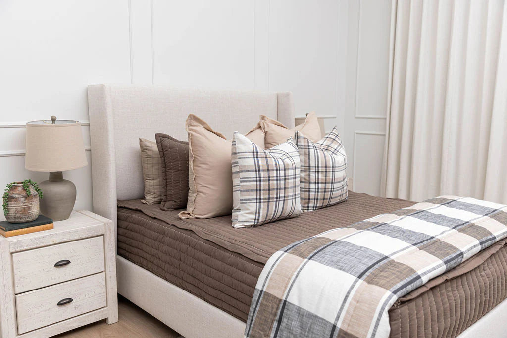Modern bedroom with a beige upholstered bed featuring Airstream RV Fit Beddy’s bedding designed for Pottery Barn Travel Trailers, styled with a brown quilt, plaid blanket, layered neutral pillows, a white nightstand, a beige lamp, a small plant, and light curtains.