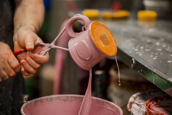 A close-up of a person's hands using a metal scraper to remove excess pink paint from the handle of a pink ceramic mug, which is balanced on its orange base over a pink paint bucket on a workshop table.