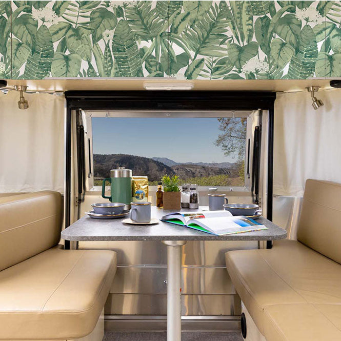Interior of a camper van with tan seats, a table set for breakfast, an open book, and a window revealing a scenic mountain view. The upper wall features Tropical Peel + Stick Removable Wallpaper with green leafy designs, adding a fresh touch to the inviting Airstream interior décor.