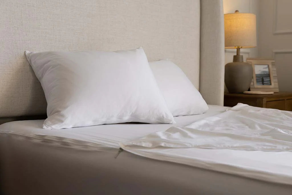 A neatly made bed, outfitted with Airstream RV Fit Beddy’s ZipBase Sheets for Flying Cloud Travel Trailers and two white pillows, sits next to a wooden nightstand with a lamp and a framed photo.