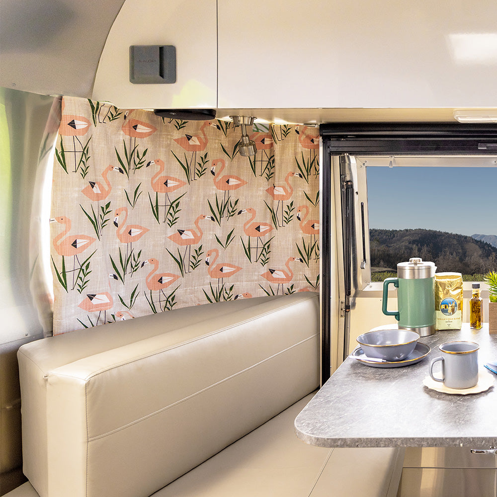 A retro RV interior with beige seating, a table set with dishes and a thermos, Airstream Printed Custom Curtains for Caravel Travel Trailers featuring a flamingo pattern, and a window view of trees and mountains.