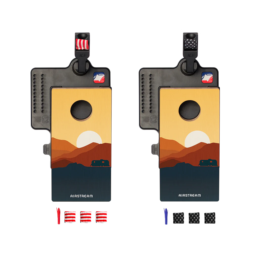 Two identical Airstream ACA Double Chuck Mini Cornhole Tabletop Game boards with sunset landscape designs and black bag hangers are shown, each with a set of red, blue, and black bean bags displayed underneath. This portable game is perfect for mini tabletop cornhole fun wherever you go.