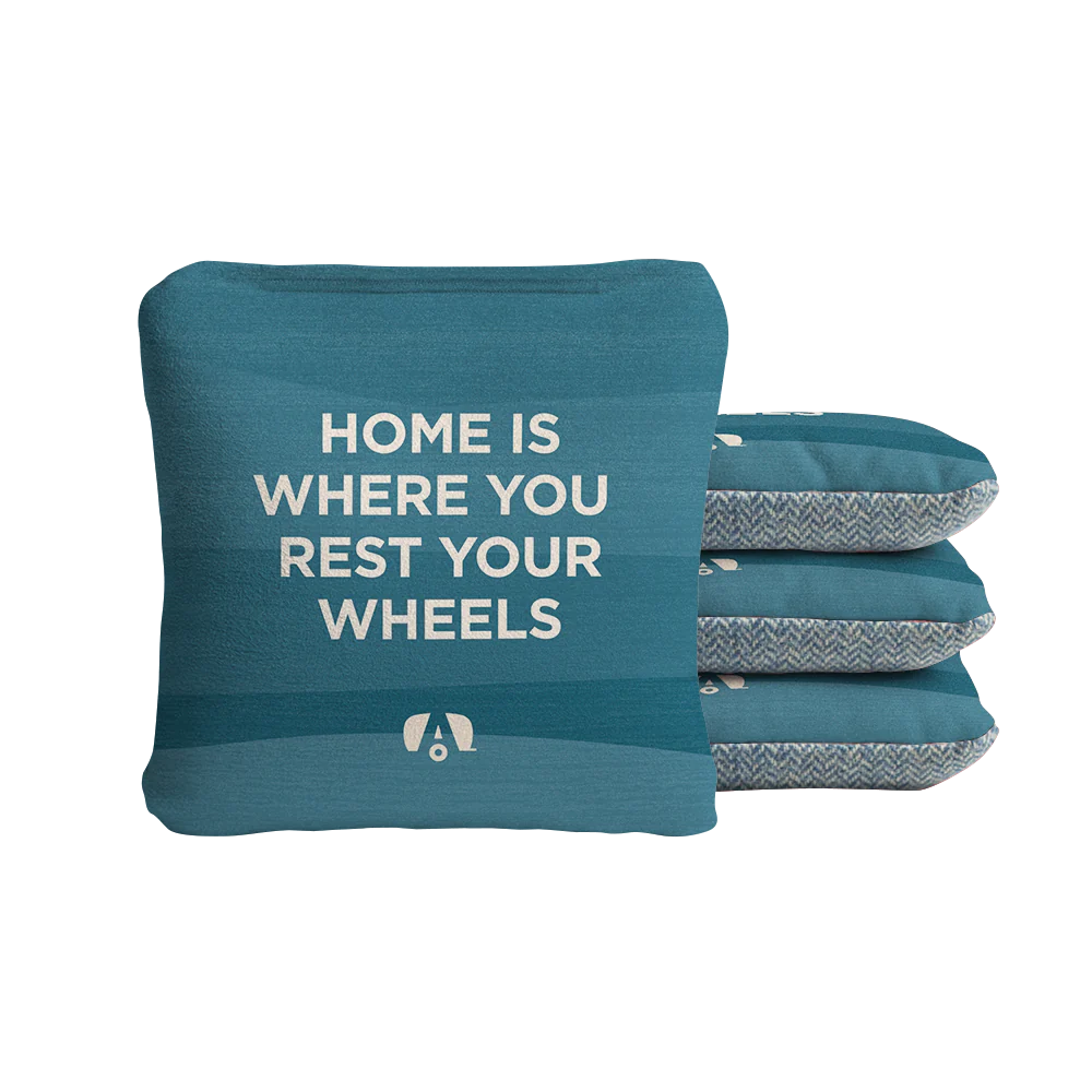 A teal throw pillow with the words HOME IS WHERE YOU REST YOUR WHEELS printed on the front, inspired by the comfort of Airstream ACA Cornhole Bags. Three more matching pillows are stacked beside it, featuring a patterned edge reminiscent of high-quality Airstream ACA Cornhole Bags.