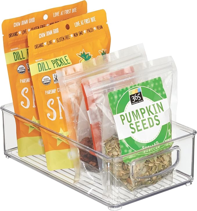 A Stackable Storage Bin (10 x 6 x 3) made of clear, BPA-free acrylic holds packs of Dill Pickle parsnip chips and a bag of shelled pumpkin seeds, all neatly arranged upright for effortless pantry organization.