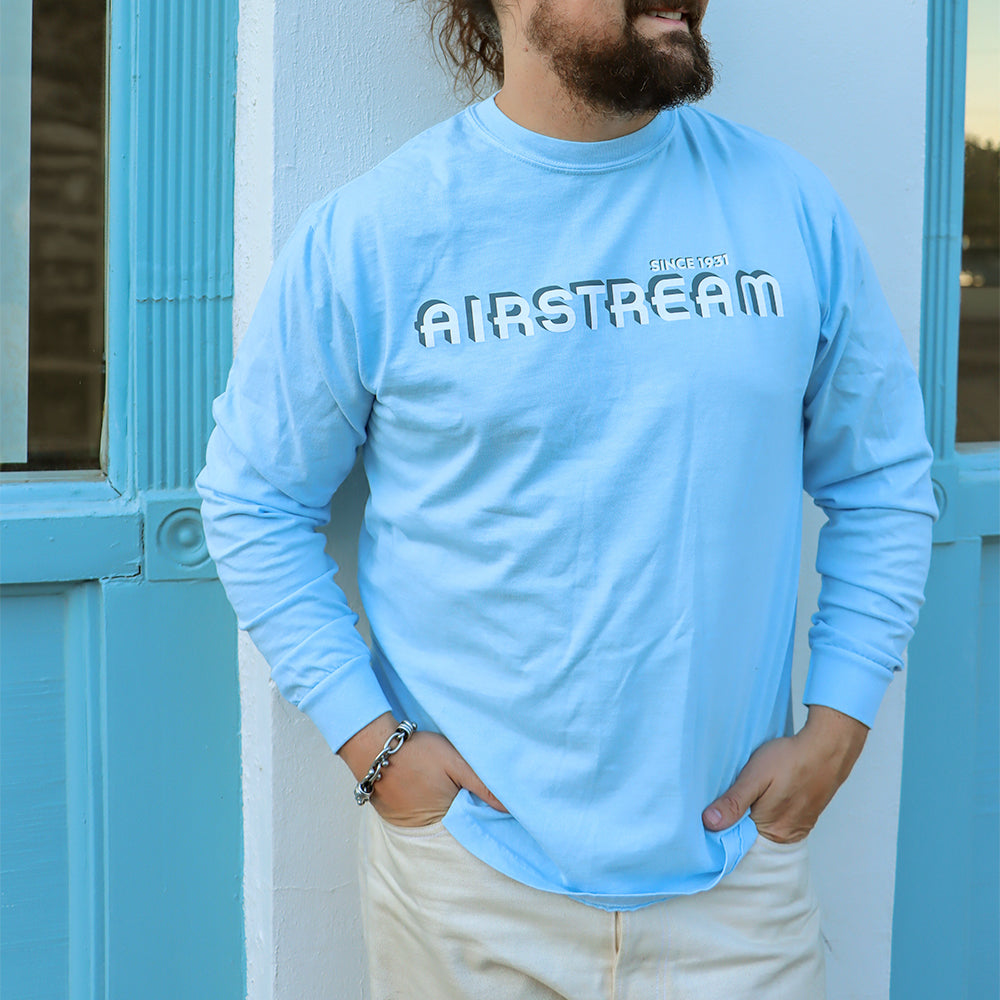 A bearded person stands in front of a blue and white building, hands tucked into the pockets of their light-colored pants, wearing the Airstream 1931 Series Long Sleeve T-Shirt in a classic light blue with “AIRSTREAM SINCE 1931” printed on it.