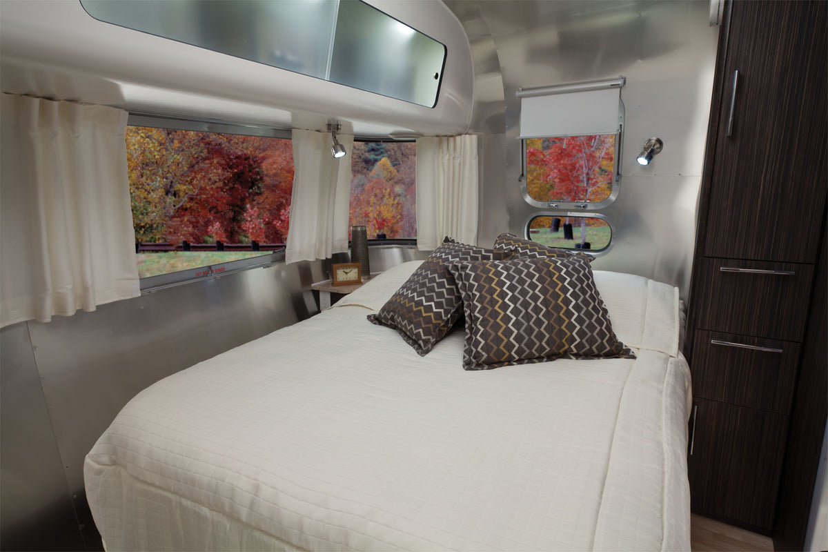 Modern RV bedroom featuring a made bed with chevron-patterned pillows, a wood side table, and large windows that showcase vibrant autumn trees outside. The cozy and minimalist interior design is enhanced by the stylish warmth and privacy provided by Airstream Solid Custom Curtains for International Signature Travel Trailers.