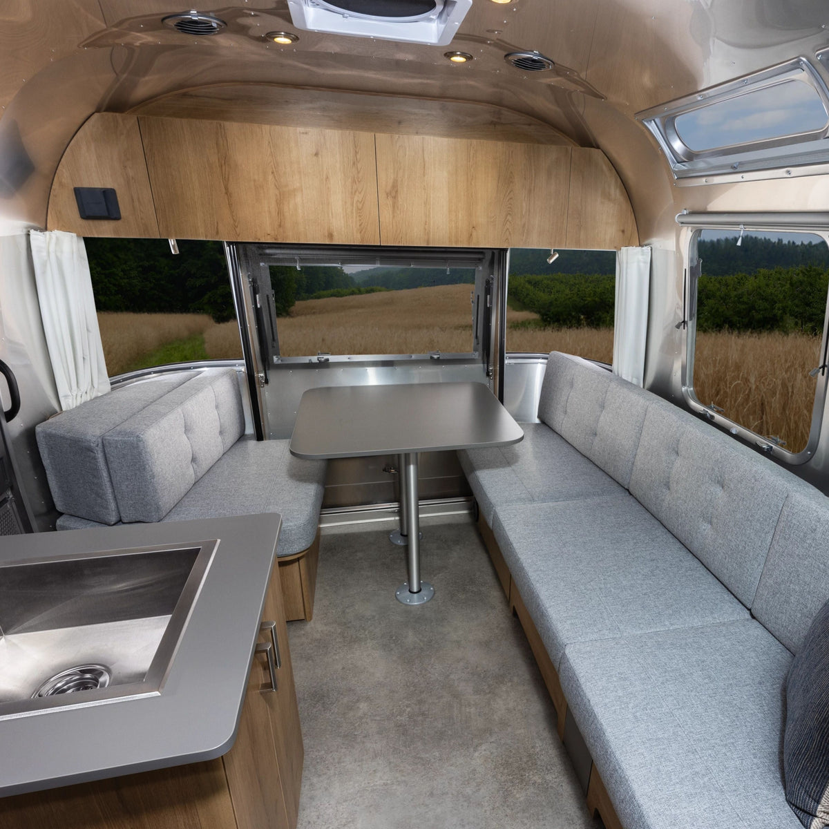Modern RV interior with light gray cushioned benches, a central dining table, wood cabinets, and a compact sink. Large windows are dressed with Airstream Solid Custom Curtains for Trade Wind Travel Trailers, offering a view of fields and greenery outside.