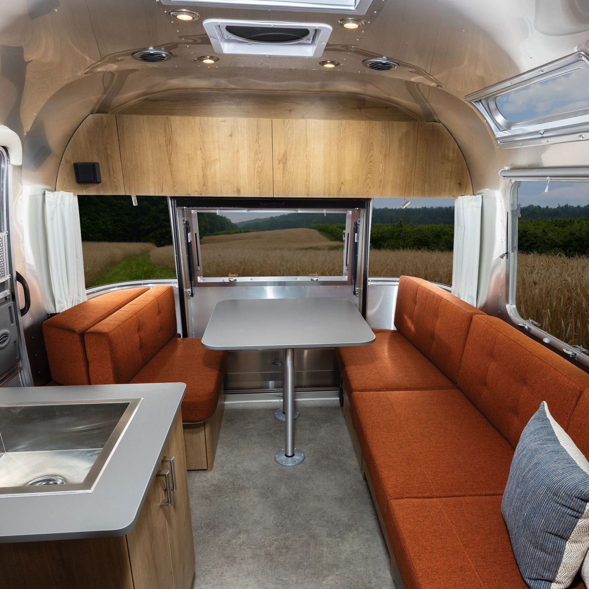 Interior of a modern RV with orange cushioned seating around a gray table, wood cabinets, a small sink, and large windows overlooking a field. The space is enhanced by Airstream Solid Custom Curtains for Trade Wind Travel Trailers, which provide privacy while complementing the contemporary design.