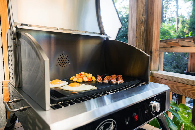 Cooking two sunny-side-up eggs, a pile of tater tots, and several pieces of bacon on the Bradley Electric Grill and Smoker, with wooden fencing and trees visible in the background.