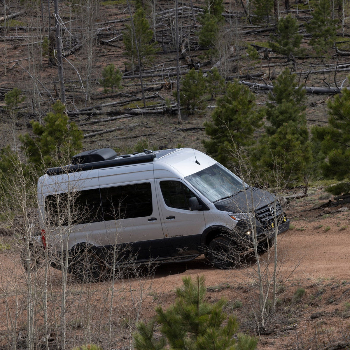 A white Airstream Interstate 19X camper van drives up a dirt road through a forested area with young green trees and patches of brown earth, the Airstream Owner Manuals: Interstate 19X spiral-bound and tucked neatly inside for the journey ahead.