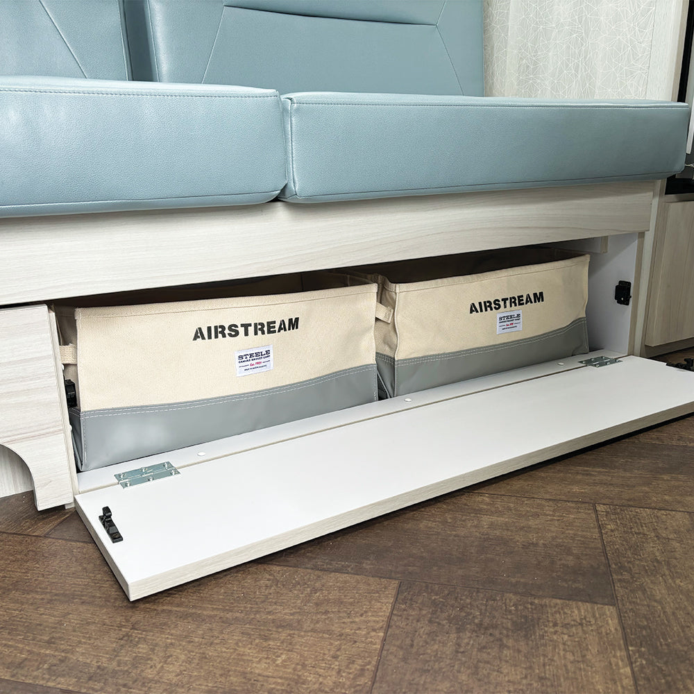 Two beige and gray Airstream x Steele Canvas 25′ and 27′ Lounge Storage Bins, labeled AIRSTREAM, are neatly stored beneath a light wood bench seat topped with a blue cushion. The front panel is open, displaying the custom canvas bins inside. The storage organizer highlights the bins arranged above a dark, herringbone-patterned floor.