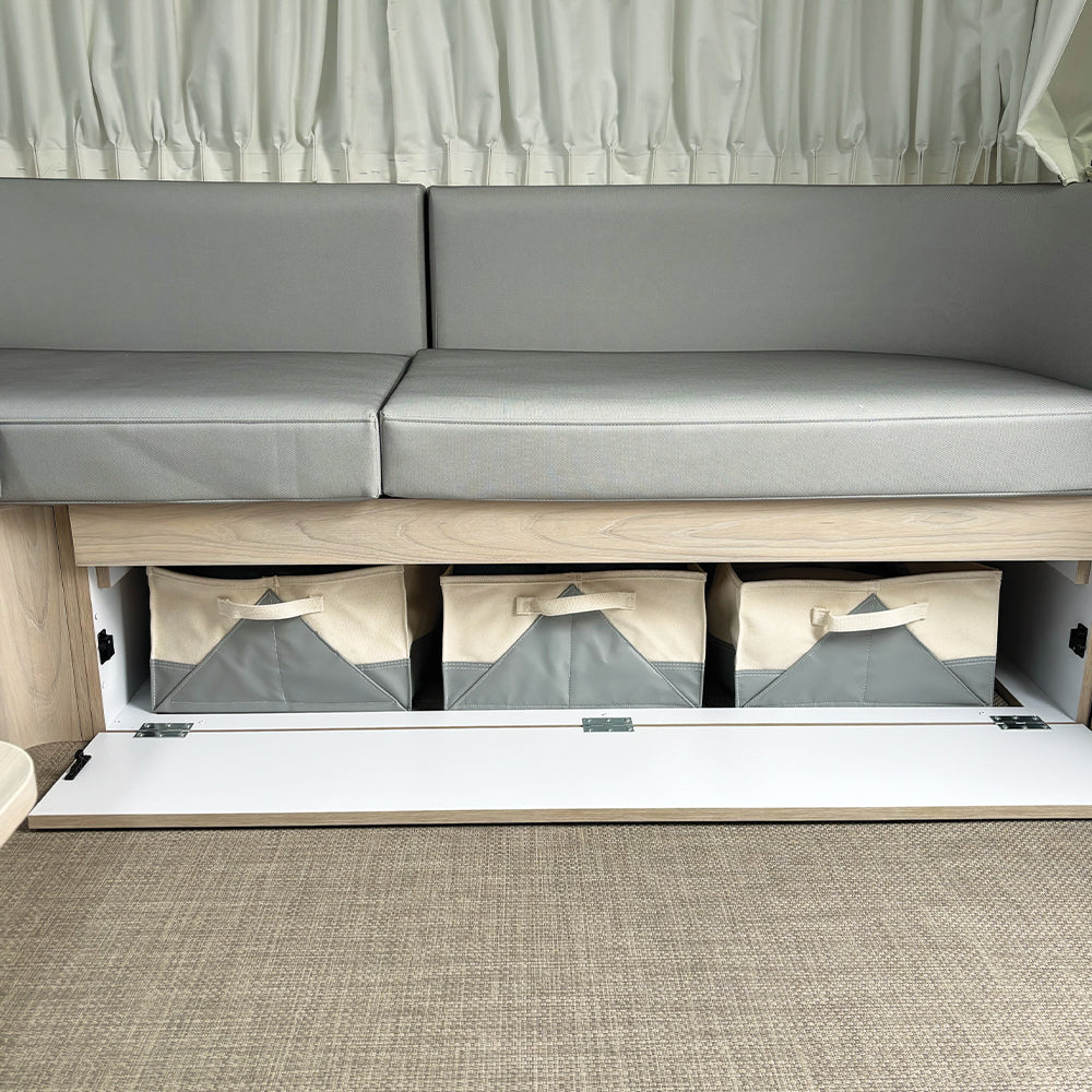 A light gray couch with a storage compartment underneath holds three Airstream x Steele Canvas 28′ and 30′ Lounge Storage Bins in beige and gray fabric, including a custom canvas bin ideal as a travel trailer organizer. The storage area features a white hinged door that opens downward. Curtains hang behind the couch, and the floor is carpeted.