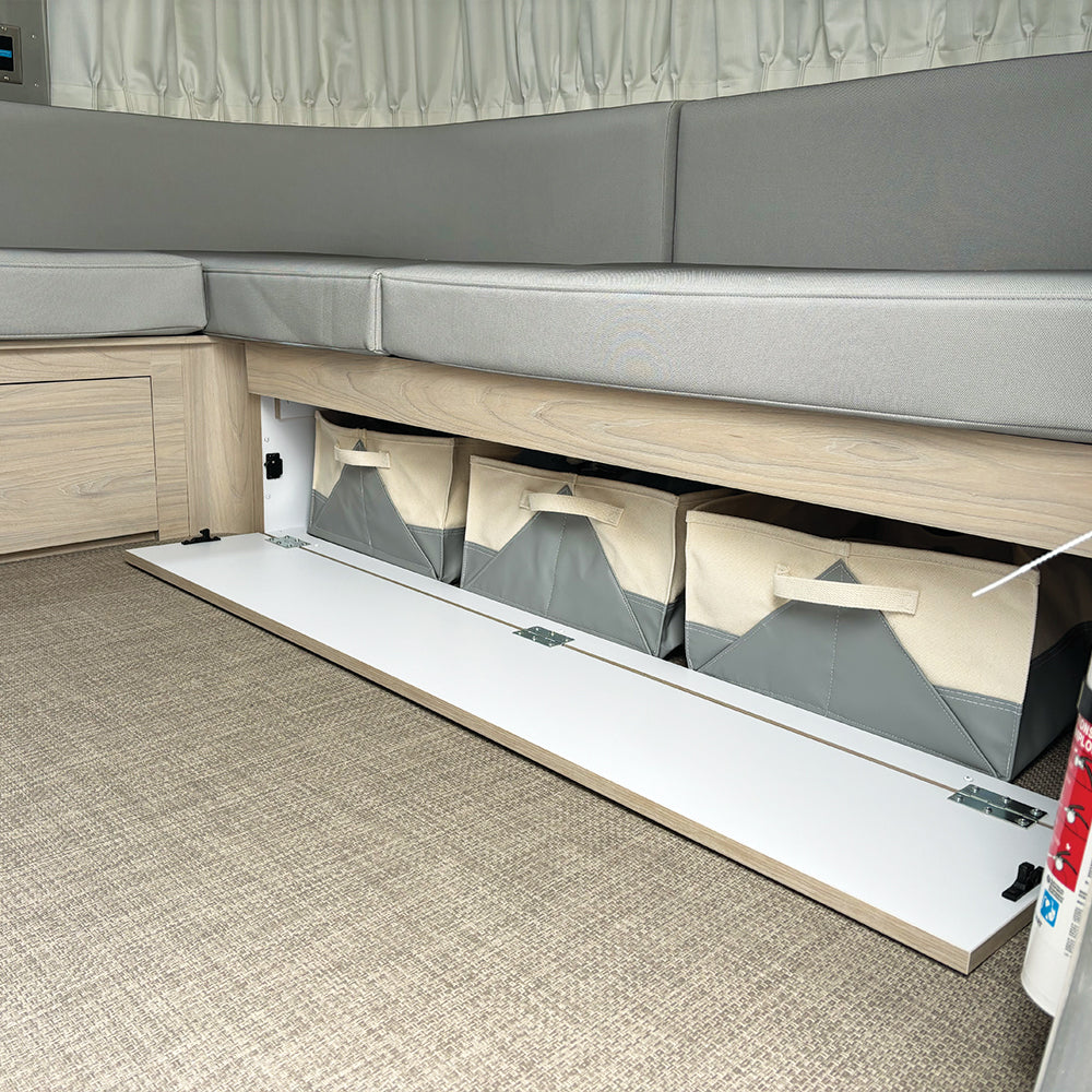 The bench seat with light gray cushions lifts to reveal under-seat storage, where three Airstream x Steele Canvas 28′ and 30′ Lounge Storage Bins are neatly organized; this area features light wood cabinetry and beige carpet flooring, creating an ideal travel trailer organizer solution.