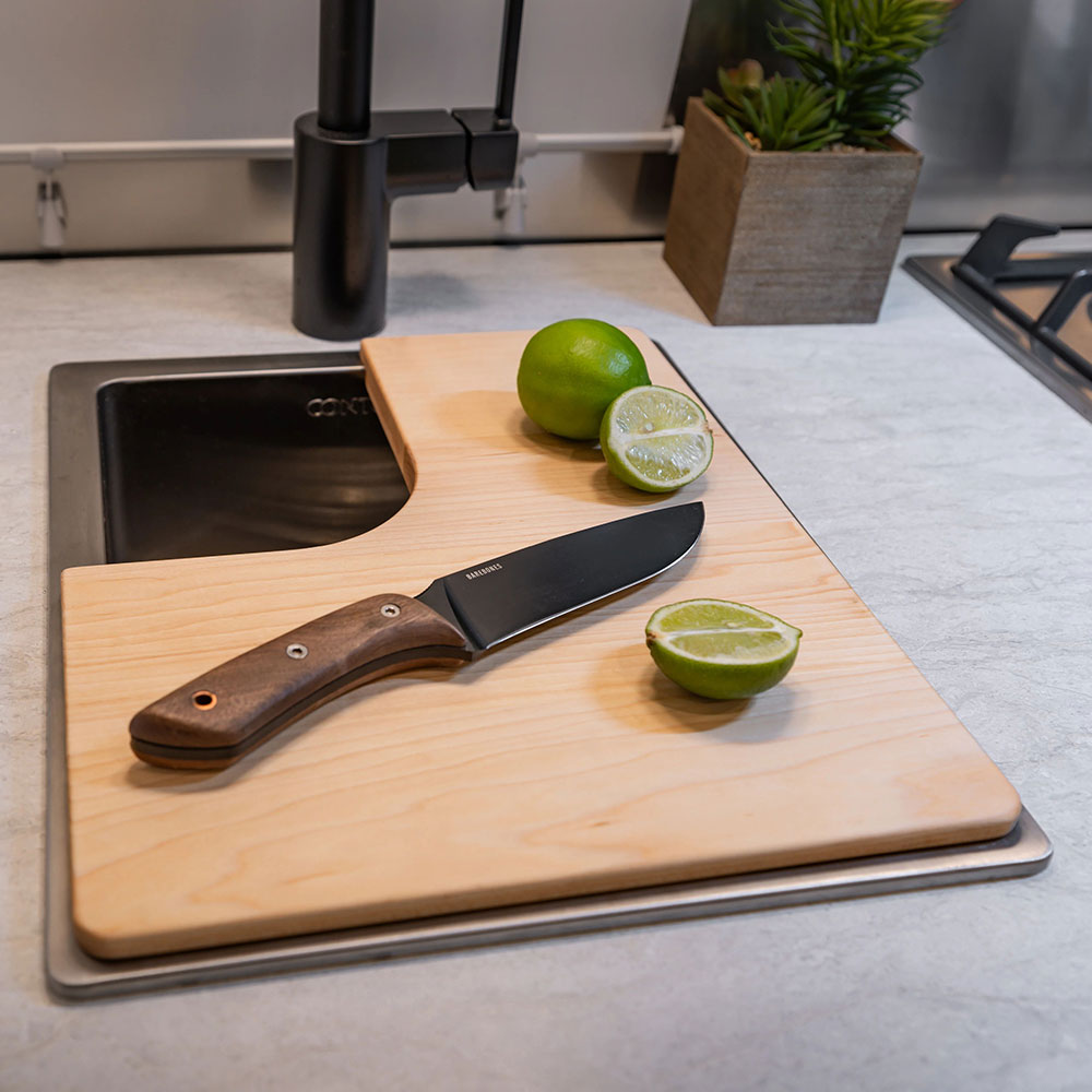 A kitchen knife rests on a wooden cutting board next to a halved lime and a whole lime, near a sink outfitted with an Airstream Custom Sink Cutting Board for Sport Travel Trailers. A small potted plant sits nearby on the countertop, adding to the practical charm of this functional space.