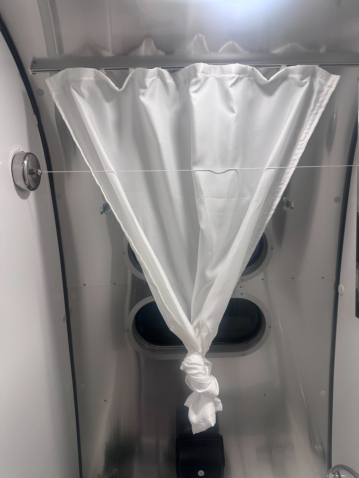 A white curtain is tied in the middle, forming a V shape, and hangs in front of two small oval windows inside a metallic-walled space—an inviting touch that demonstrates how the Airstream Retractable Interior Clothesline (Part # 382063) can bring both function and style to your Airstream.