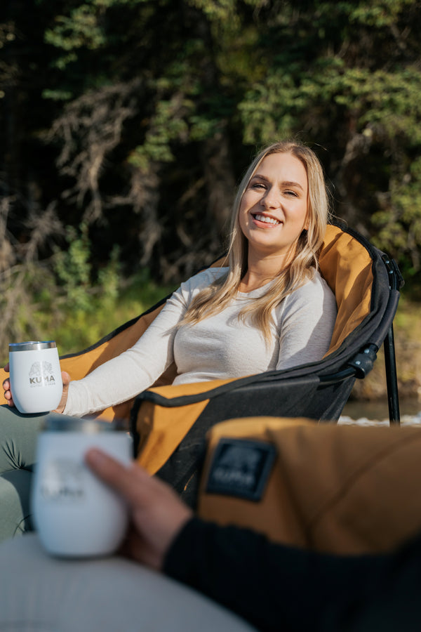 A woman sits in a black and tan camping chair outdoors, smiling while holding a white insulated tumbler. Another person in the foreground holds a matching tumbler.