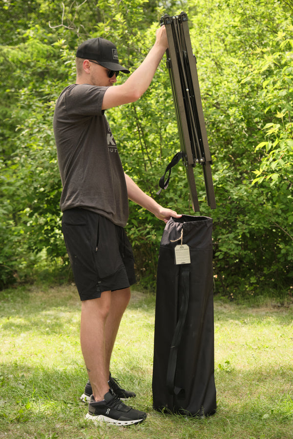 A man outdoors on grass wearing a black cap and sunglasses, holding a folded dark green folding table next to its tall black carrying bag.