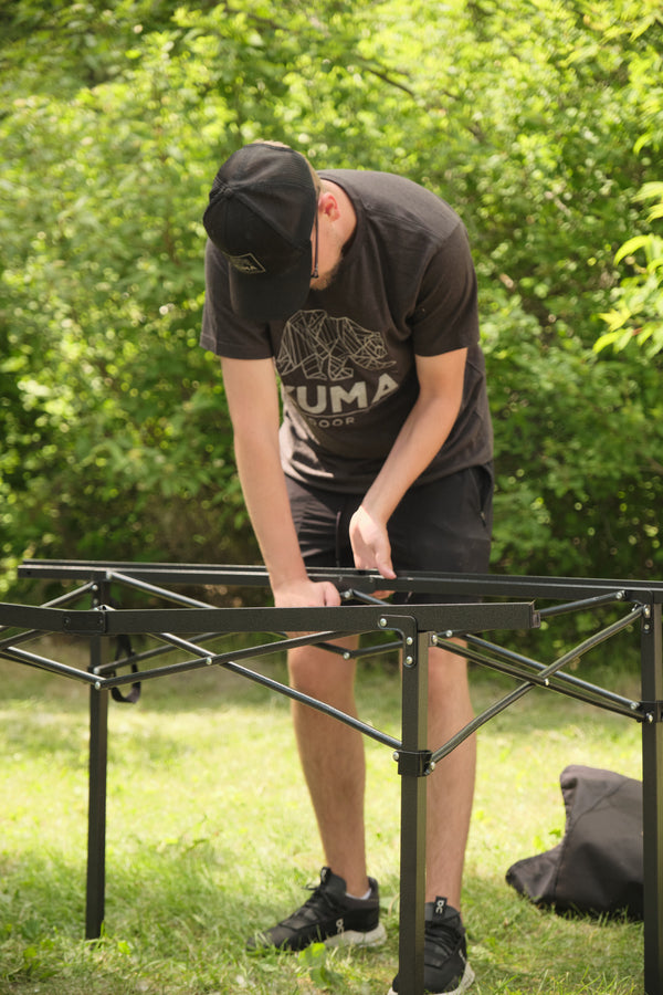 A man wearing a black Puma t-shirt and shorts is assembling a black metal folding table outdoors on a grassy lawn with trees in the background.