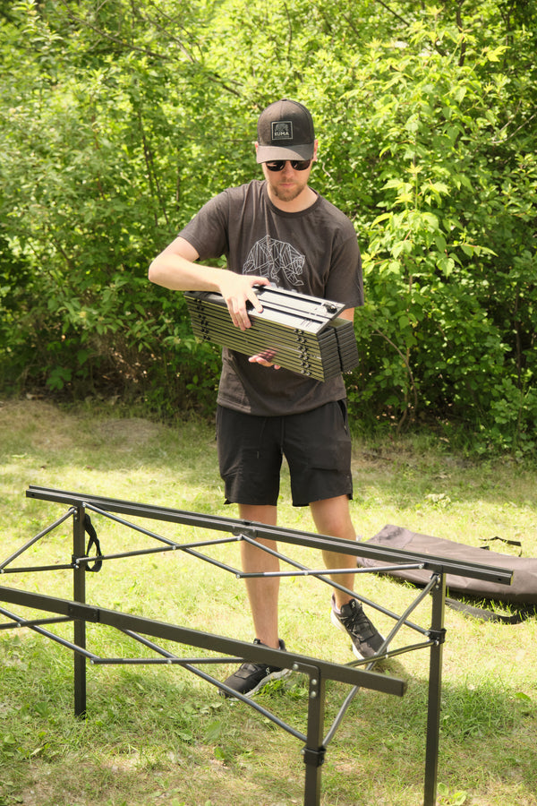 A man outdoors on grass holding a folded metal camping table frame, with a larger assembled black metal table structure in front of him and leafy green trees in the background.