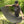 A man outdoors on grass in front of dense green trees is demonstrating a curved black slatted lounge chair or fire pit cover resting on a metal frame. He is wearing a black Kuma t-shirt and a black baseball cap.