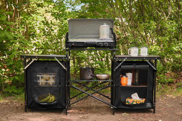 A black outdoor cooking and storage system set up in a wooded campsite environment. The system includes a built-in grill on top, a central folding table, and two side storage cabinets with mesh doors, filled with camping gear and food.