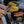 A close-up action shot of a person using metal tongs to assemble a hot dog with caramelized onions, with a cast iron skillet containing hot dogs and onions on a portable stove in the background.