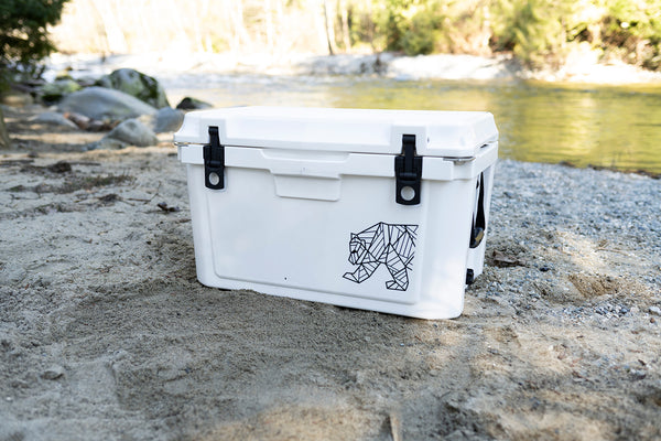 A white cooler with black latches and a black geometric animal graphic, sitting on a sandy riverbank with water and trees in the background.