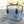 A light gray cooler with a black handle and latches, sitting on sandy ground outdoors near water and trees. A geometric bear logo is printed on the front.