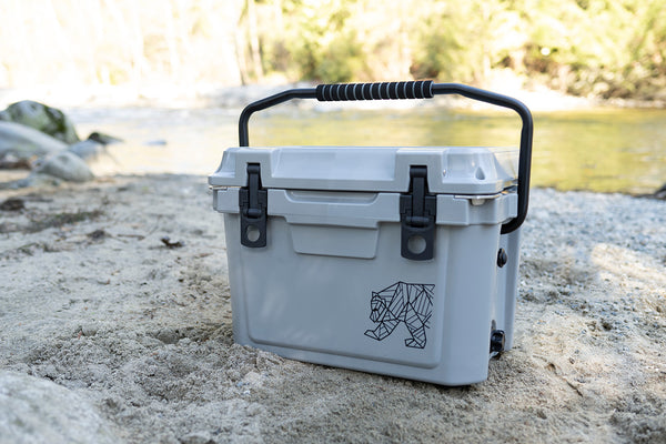 A light gray cooler with a black handle and latches, sitting on sandy ground outdoors near water and trees. A geometric bear logo is printed on the front.