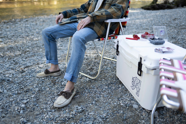 A person sitting outdoors on a folding camp chair beside a white cooler on a gravel surface near water. The person wears a plaid shirt, light blue jeans, and loafers, and the cooler has a small printed logo on the side.