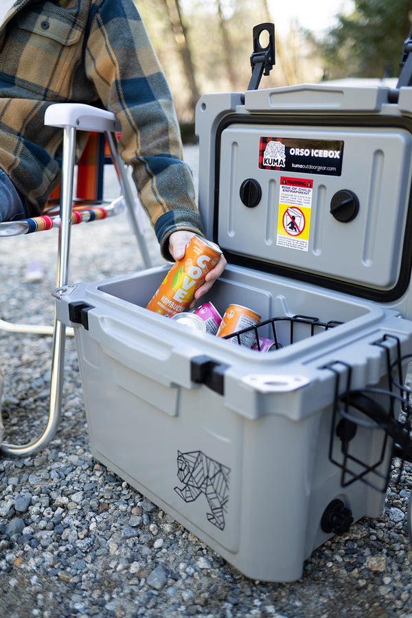 A gray Osco Icebox cooler sitting outdoors on gravel, with a person in a plaid shirt placing an orange can into the cooler basket.