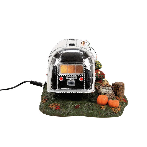 A decorative miniature silver trailer or camper on a textured green base with small pumpkins, a tree stump, and autumn leaves, shown on a plain white background with a black power cord extending to the left.