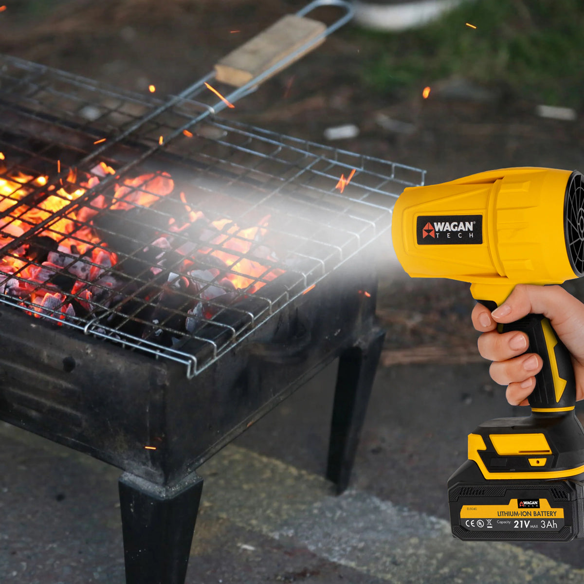 A person uses the Jetblow by Wagan Tech, a yellow compact blower, to stoke a charcoal grill with glowing coals, increasing airflow for a hotter fire.