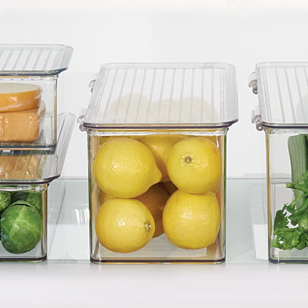 Clear plastic food storage containers with ribbed lids, shown on a refrigerator shelf. The center container holds several yellow lemons, with other containers partially visible holding sliced orange fruit, green Brussels sprouts, and green herbs.
