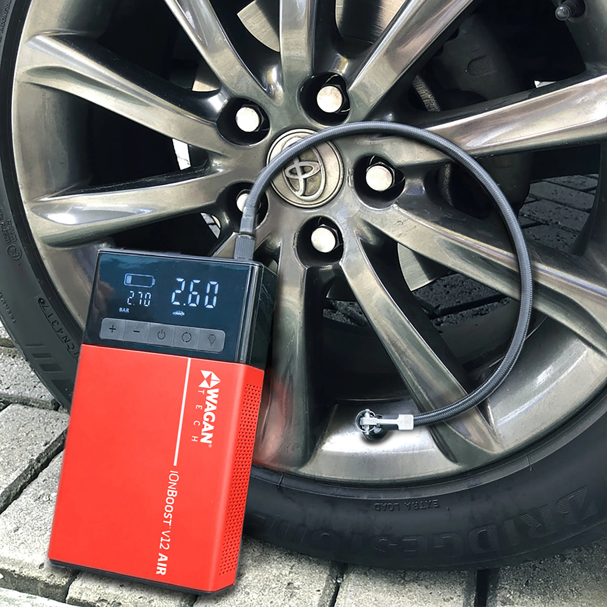 The iOnBoost™ V12 Air Compressor/Jump Starter by Wagan Tech is inflating a car tire in this scene. This versatile device, which features a digital display showing real-time pressure readings, is connected securely to the tire’s valve. The car wheel bears the Toyota logo. For added convenience on the road, the iOnBoost™ V12 also functions as both a portable power bank and a jump starter.
